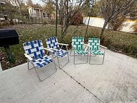 Four folding lawn chairs outdoors on concrete patio: two with blue and white woven webbing, and two with green and white woven webbing.