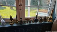 Row of various antique and vintage bells on a window ledge including brass, cast iron, and glass bells in different styles and sizes.