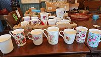 Overall view of approximately 15 bone china mugs with floral and botanical designs arranged on a wooden surface showing diverse styles and colors.