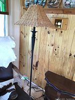 Floor lamp standing next to wooden wall with wicker shade and black metal base visible