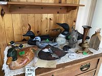 Photo showing multiple bird figurines including duck decoys, a loon, metal bird figure, and a metal vase on a cloth-covered table against a wood panel wall