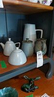 Set of pottery items displayed on shelf including white sugar bowl with lid, white creamer with lid, ceramic carafe, small jar, and teapot