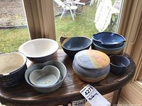 Image showing nine signed pottery pieces including bowls and one lidded bowl displayed on a wooden bench by window with outdoor view.