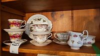 All items arranged on wooden shelf showing five teacups and saucers, December cake plate in the back, cream and sugar set on the right.