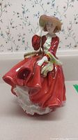 Front angle view of Royal Doulton figurine dressed in red with hat and green scarf, detailed facial features and ruffled dress.