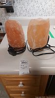 Pair of orange-pink Himalayan salt rock lamps on black bases with electrical cords placed on a white countertop, with product tag visible.