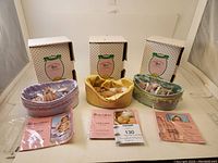 Three Paradise Galleries Treasure Collection dolls with their original boxes and paperwork displayed side by side. Each doll has a different colored basket or laying nest (purple, yellow, green).