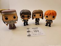 Front view of four Funko Pop Harry Potter figures: Albus Dumbledore, Harry Potter, Hermione Granger, Ron Weasley. Figures standing on flat surface with a note showing lot number and description.