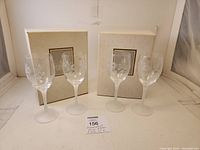 Four clear glass wine glasses with etched hummingbird designs standing in front of two Avon branded boxes with a MaxSold lot card in front.