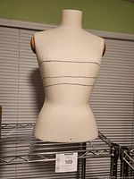 Front view of the fabric sewing bust mannequin on a metal wire shelf, showing the overall shape and fabric covering with marked horizontal lines.
