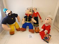 Four plush dolls of Popeye characters displayed: Popeye, Brutus, Olive Oyl, and Sweet Pea. Popeye and Sweet Pea have tags.
