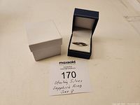 Ring displayed inside its blue cushioned box with outer white box beside it and a lot card in front.