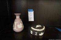 Lavender jasperware bud vase with white relief decoration and black jasperware oval dish with lid shown on a shelf.