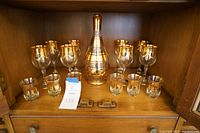 Full view of wine glasses, shot glasses, and decanter with gold detailing inside wooden cabinet.