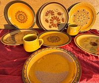 Photo of seven vintage ceramic plates and two yellow cups arranged on a red cloth, showing the front design details of the plates and cups.
