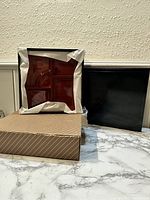 Two Japanese bento box trays visible, one partially wrapped in tissue paper, displayed upright next to their black outer box with striped cardboard sleeve on marble surface under indoor lighting.