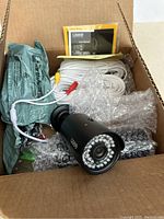 Top view of the open box containing one black QSEE bullet-style security camera with attached cables, manual, protective packaging, and bubble wrap.