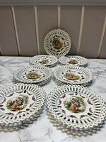 Set of six white porcelain trinket dishes with pierced lattice edges arranged on a marble surface, each with a colorful Victorian couple scene and floral detail