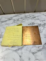 Photo of the brass printer plate and accompanying yellow paper with printed wedding invitation text on a marble surface.