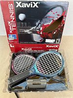 Front view of sealed XaviX Tennis game box including two tennis racket controllers and accessories inside clear plastic packaging.