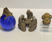 The three Asianware pieces displayed together: a blue glass paperweight with a brass goldfish, a mudmen figurine trio playing Chinese chess, and a small resin figurine seated with arms behind head.