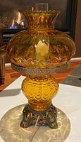 Side view of the amber glass hurricane lamp with lit bulb inside, showing illuminated glass and ornate metal base.