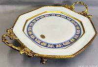 Top angled view of the octagonal porcelain dish showing blue and floral pattern with gold rim and ornate metal handles and feet.