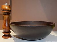 Photo of dark wooden pepper mill standing next to large dark wooden salad bowl on plain surface with brown background.