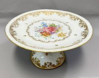 Angled front view of white porcelain cake stand with floral hand-painted bouquet in center and gold ornate trim around edge and base.