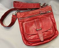Front view of red leather shoulder bag with flap pocket, zippered pocket above, and adjustable strap laid flat.