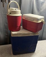 Photo shows a large blue Coleman cooler, a medium red rectangular Coleman cooler with beige lid and latch, and a red and beige Coleman thermos with handle arranged on a surface.
