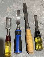 Four assorted woodworking chisels with different sized steel blades and various plastic and wood handles laid out on a concrete floor.