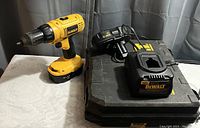 Yellow and black cordless drill with battery attached, battery charger, displayed on and next to a table, grey curtain background