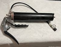 Photo of a black handheld grease gun tool with metal lever and attached hose on a flat surface.