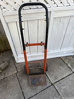 Front view of a metal hand held dolly with black handle and orange frame placed on concrete ground in front of a white fence.
