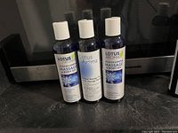 Three dark blue bottles of Lotus Aroma massage and body oil set together, unopened with white caps and labels, stacked in front of a microwave.