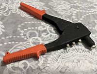 Photo of a handheld rivet tool with black metal body and bright orange plastic handles, placed on a patterned surface.