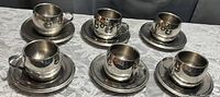 Six stainless steel Demi Tasse cups with matching saucers arranged on a tablecloth, showing polished finish and cup handles.
