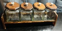 Four square glass canisters with hexagonal wooden lids, each having a white ceramic knob. All canisters are placed in a wooden stand with side supports.