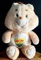 Photo of vintage plush Care Bear sitting upright with two smiling sunflowers embroidered on its belly, heart-shaped paw pads on the feet, light beige plush, brown embroidered eyebrows, and fabric nose.