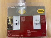 Photo of retail packaging showing two white Lutron dimmer switches with sliding controls, model details and wattage ratings visible.