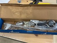 Photo of Alsons Victorian spray shower arc bar and hose inside original blue box packaging on a wooden table; items include showerhead, hose, arc bar, and mounting hardware wrapped in plastic