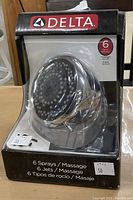 Photo of Delta shower head in original packaging showing brand logo, product visible through plastic with multiple nozzles, and printed information about 6 sprays and massage.