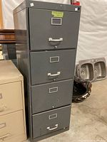 Front and angled view of the gray four drawer filing cabinet showing metal construction, handles, and label holders on each drawer. Used with scratches and one drawer not fully closed.