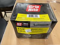 Box of Grip Rite masonry nails on a wooden table, showing label with size 3-1/2 inch, weight 1.5 kg, fluted tempered hardened steel.