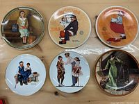 Photo showing six Norman Rockwell collector plates arranged in two rows of three on a flat surface. Each plate features different vintage Norman Rockwell art scenes, including children and adults in period clothing and some plates referencing Christmas.