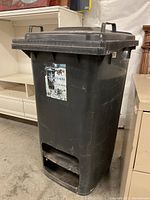 Full view of the black plastic salt bin with lid and handles, showing wear and a torn label on the front. The bottom opening is visible for dispensing salt.