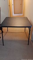 Full view of the black square card table set up in a hallway, showing the top surface and legs.