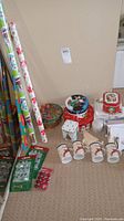 Photo showing rolls of Christmas wrapping paper standing vertically with festive patterns and various Christmas tins, one square and multiple round tins with holiday motifs next to four ceramic mugs with Christmas designs.