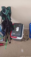 Folded green camp chair, Koolatron plug-in cooler in white basket, and other containers on carpeted floor.